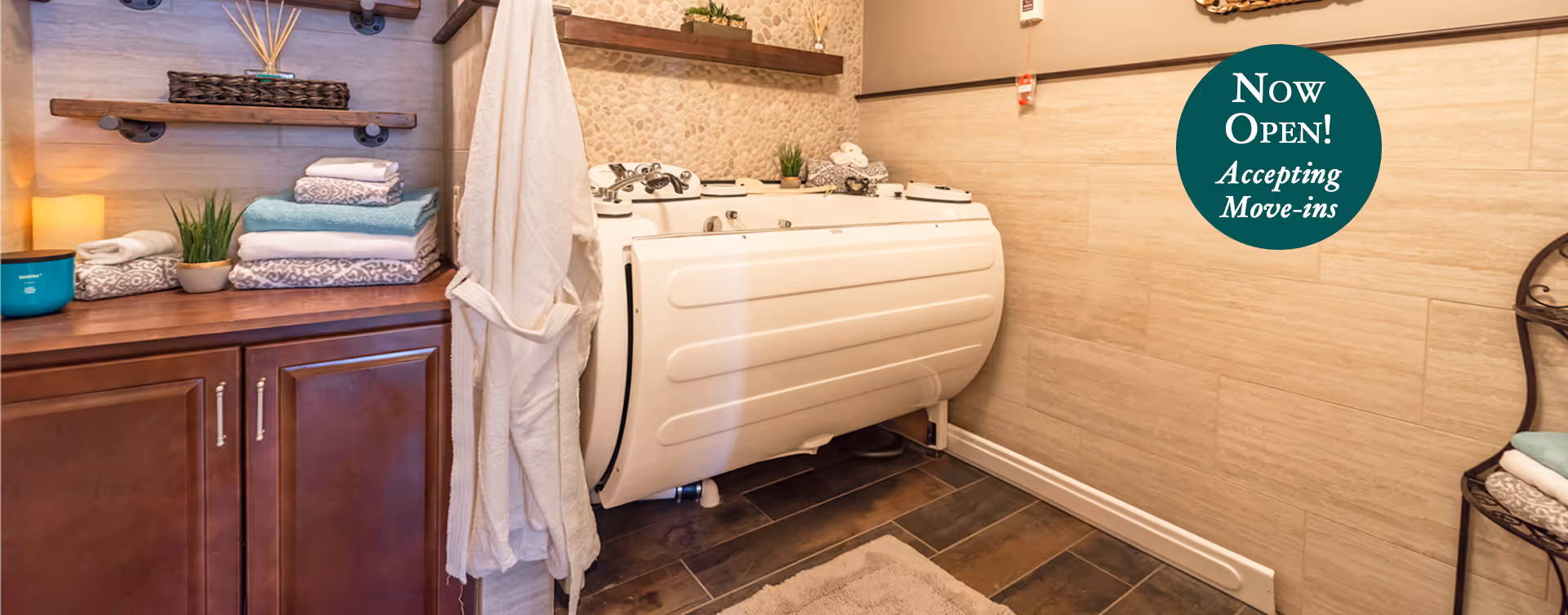 A bathroom with a walk-in bathtub, wooden cabinets, folded towels, a white bathrobe hanging on a hook, and decorative plants. The walls have a mix of stone and tile finishes, and there is a small rug on the floor. A green circular sign on the wall reads 'Now Open! Accepting Move-ins'.