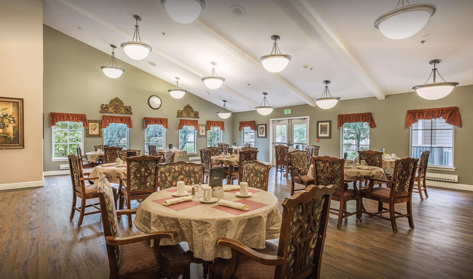 A spacious dining room with multiple round tables covered with beige tablecloths, each set with cups, napkins, and condiments. The room has wooden chairs with floral upholstery, green walls, several windows with red valances, and ceiling lights providing warm illumination.