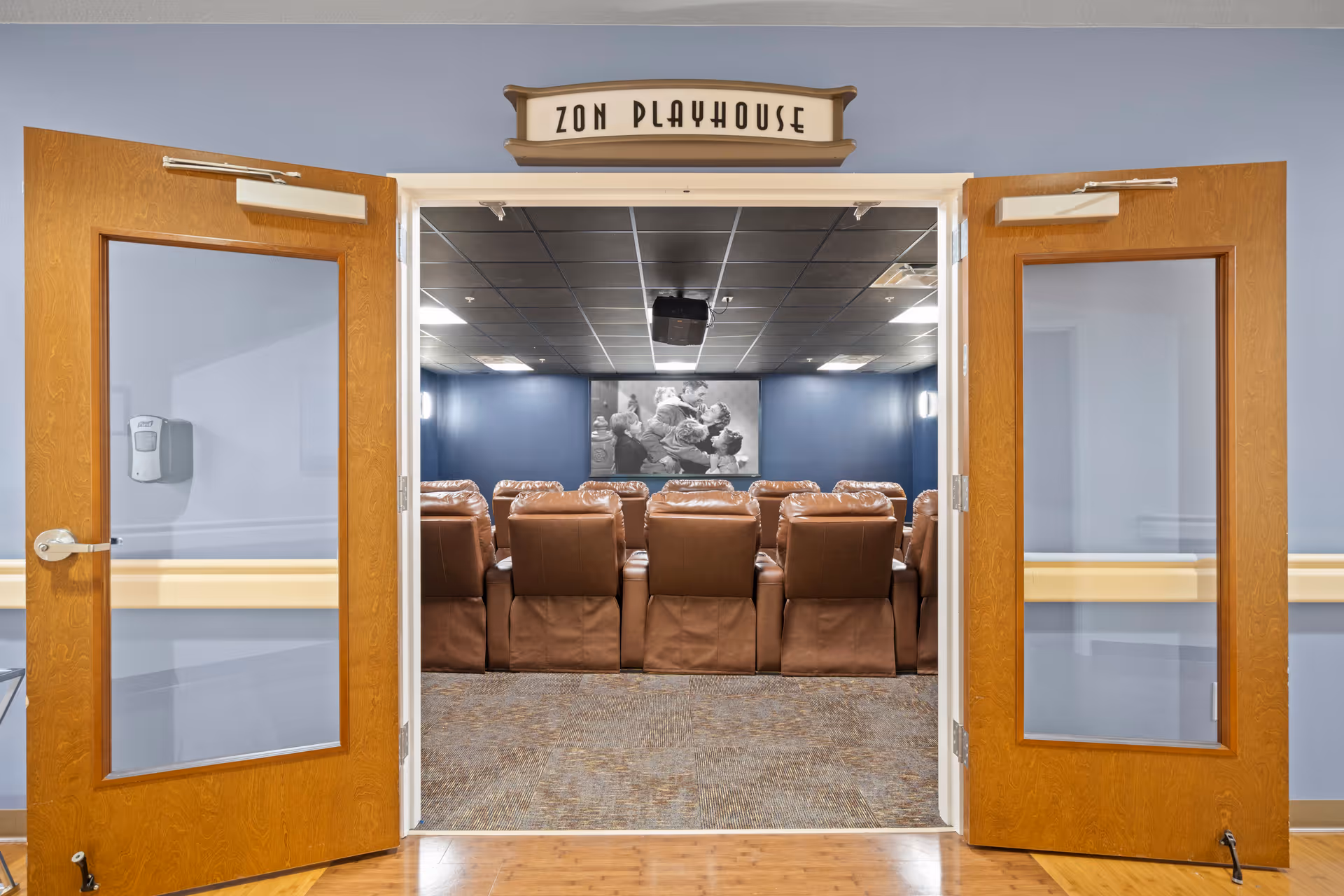 View through open double wooden doors into a small theater room with rows of brown leather recliner chairs facing a screen. Above the doors is a sign that reads 'ZON PLAYHOUSE'. The walls are painted blue and the ceiling has black tiles with recessed lighting.