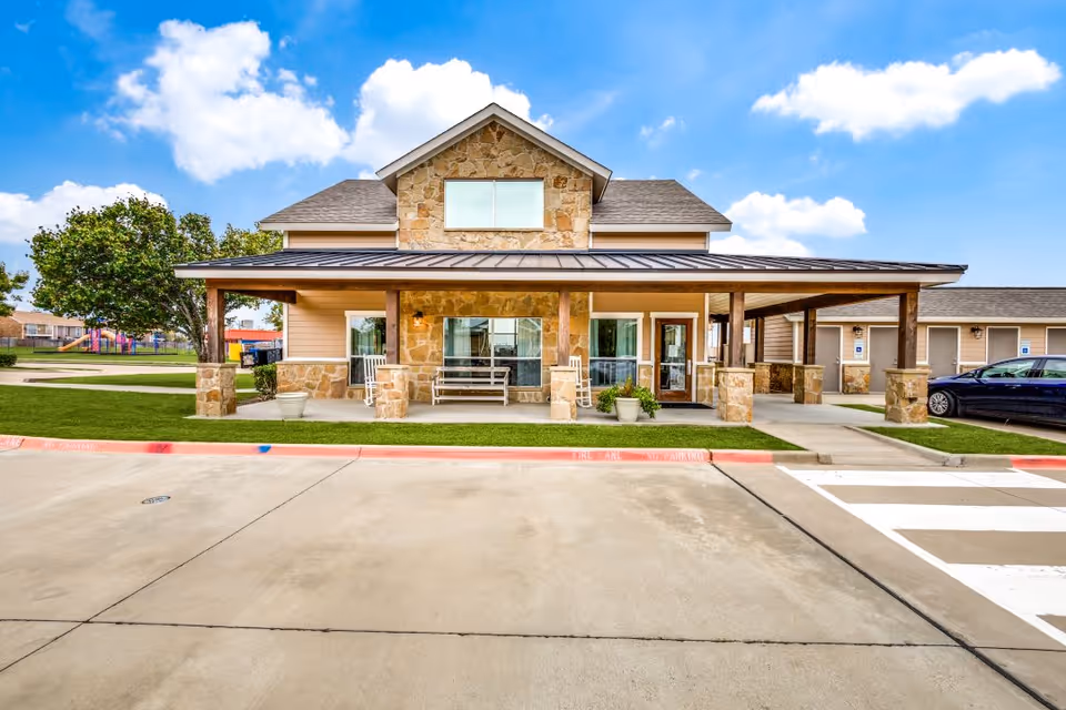 Front exterior view of a single-story senior living facility building with stone and wood siding, a covered porch with benches and rocking chairs, a parking area with marked spaces, and a clear blue sky with some clouds.