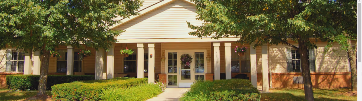 Front exterior view of Addington Place of Northville building with a covered entrance, glass double doors, beige siding, brick accents, and surrounded by green bushes and trees.