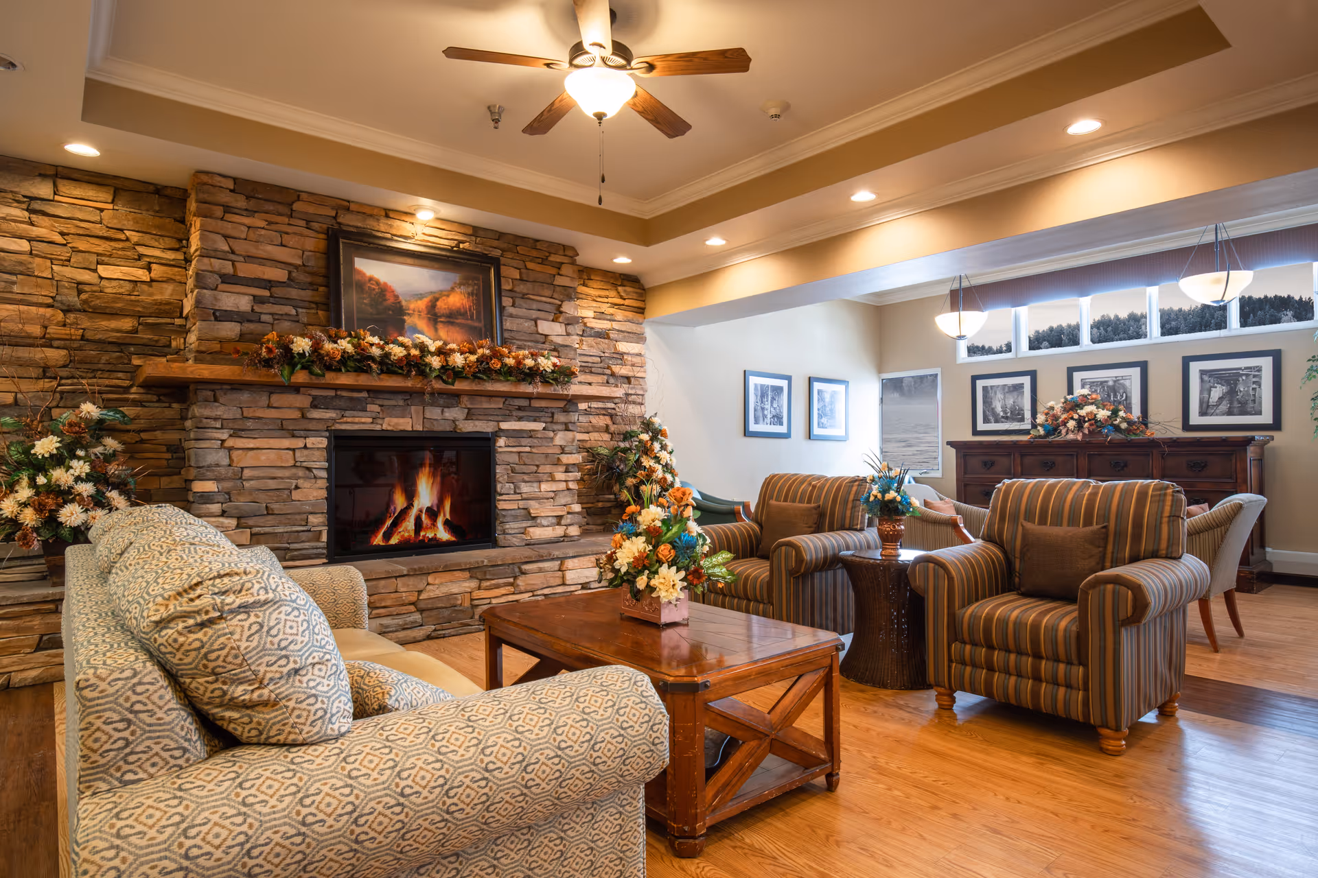 Comfortable communal living room with a stone fireplace, sofas and armchairs arranged around a wooden coffee table and floral decorations.