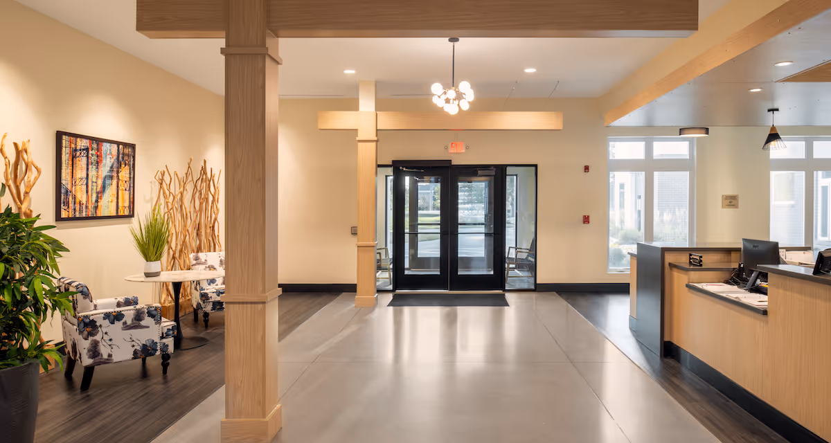 Bright and spacious lobby area with light wood accents, a reception desk on the right, double glass entrance doors straight ahead, and a small seating area with two floral armchairs and a round table on the left. There are decorative plants and a colorful framed artwork on the left wall.