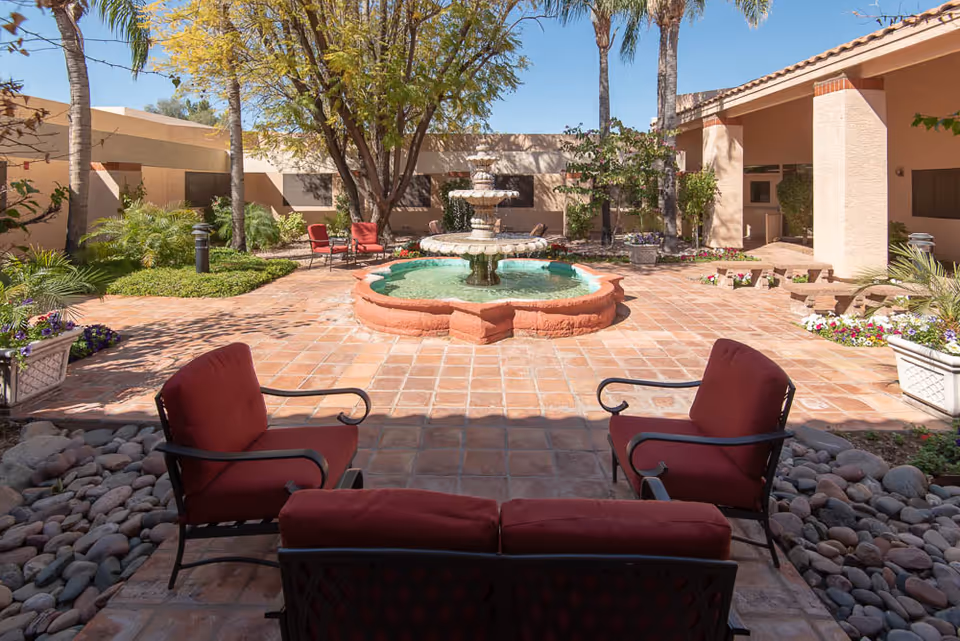 Outdoor courtyard area with a central tiered water fountain surrounded by tiled flooring. There are red cushioned chairs and a bench arranged around the fountain, with trees, plants, and flowers adding greenery to the space. The courtyard is enclosed by beige building walls under a clear blue sky.