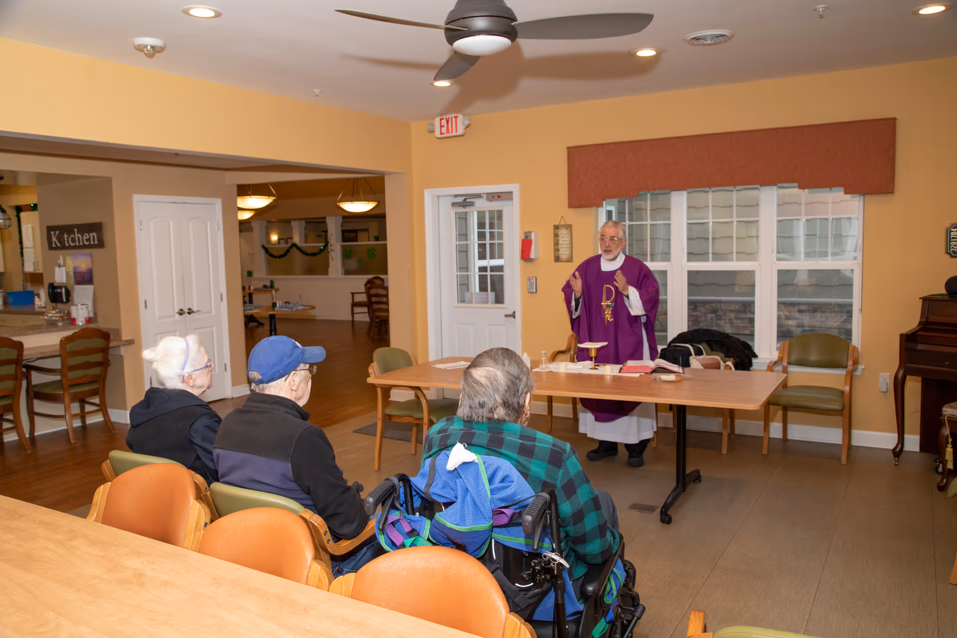 A small group of elderly people seated and facing a priest who is standing behind a table conducting a religious service in a room with yellow walls, wooden floors, and large windows. The room has chairs, a piano, and a sign indicating the kitchen area.