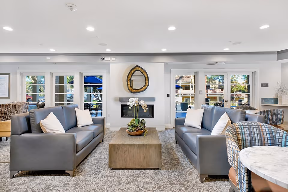 A modern living room with two gray sofas facing each other, each with white pillows. A rectangular wooden coffee table with a plant centerpiece is between the sofas. Behind the coffee table is a fireplace with a decorative mirror above it. Large windows and glass doors let in natural light and show an outdoor patio area with blue umbrellas. The room has recessed ceiling lights and patterned carpet flooring.