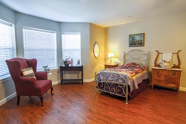 A cozy bedroom with a single metal bed featuring a colorful quilt, a wooden nightstand with a lamp, a wooden dresser with decorative items, a red upholstered armchair with a pillow, and a small table with flowers in front of three windows with blinds.