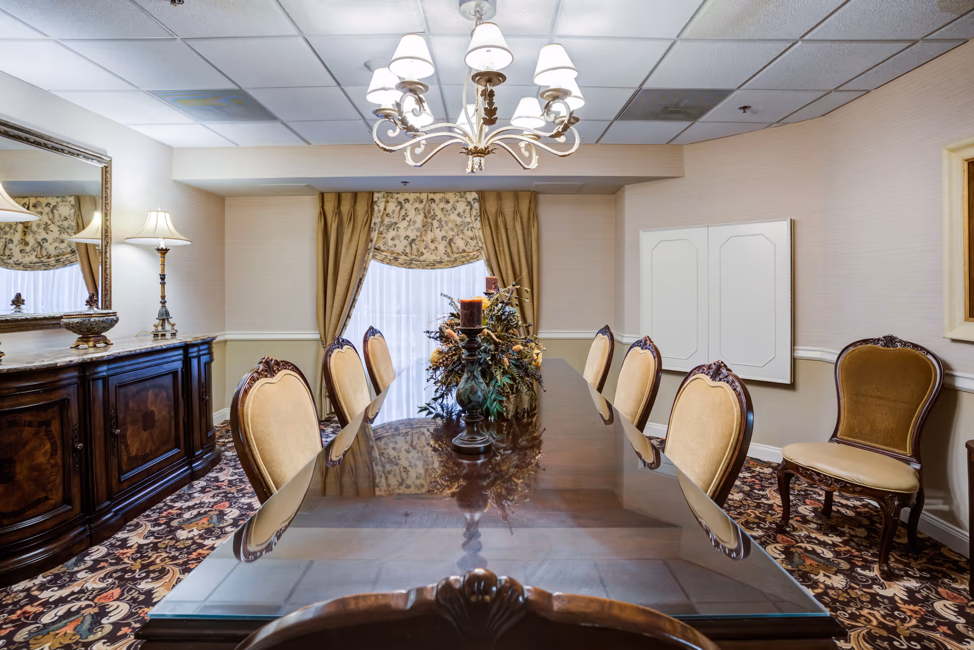 A formal dining room with a long polished wooden table surrounded by eight upholstered chairs. A decorative centerpiece with candles is placed in the middle of the table. The room features patterned carpet, beige walls with a chair rail, a large mirror above a dark wooden sideboard, a floor lamp, and a chandelier hanging from the ceiling. A window with floral curtains and drapes is at the far end of the room.