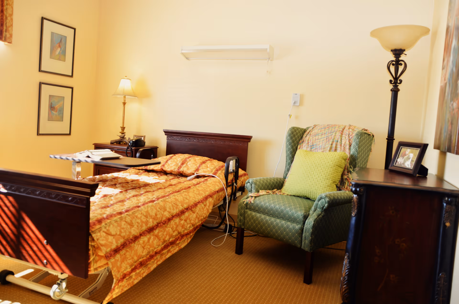 Furnished senior living bedroom with a hospital-style bed, bedside tables, lamps, and a green upholstered armchair.