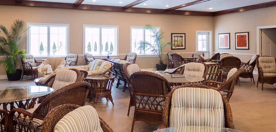 A spacious senior living common area with multiple wicker chairs and glass-top tables arranged throughout the room. The room has large windows letting in natural light, potted plants, and framed artwork on the walls. The ceiling features wooden beams and recessed lighting.