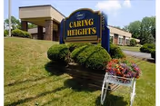 Outdoor view of Caring Heights Community Care and Rehabilitation Center sign on a grassy area with trimmed bushes and a flower planter in front. The building and driveway are visible in the background under a partly cloudy sky.