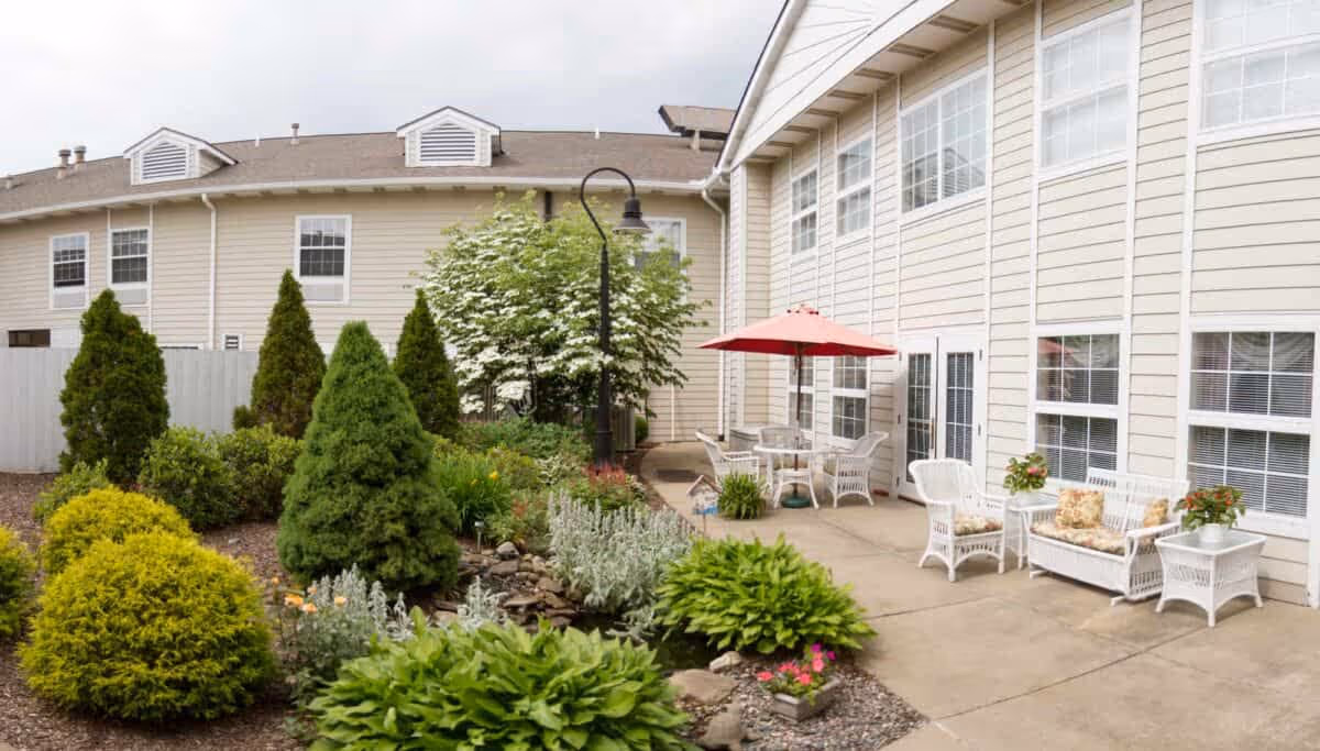Outdoor courtyard with wicker chairs and tables under a red umbrella beside landscaped shrubs and the beige exterior of the senior living building.