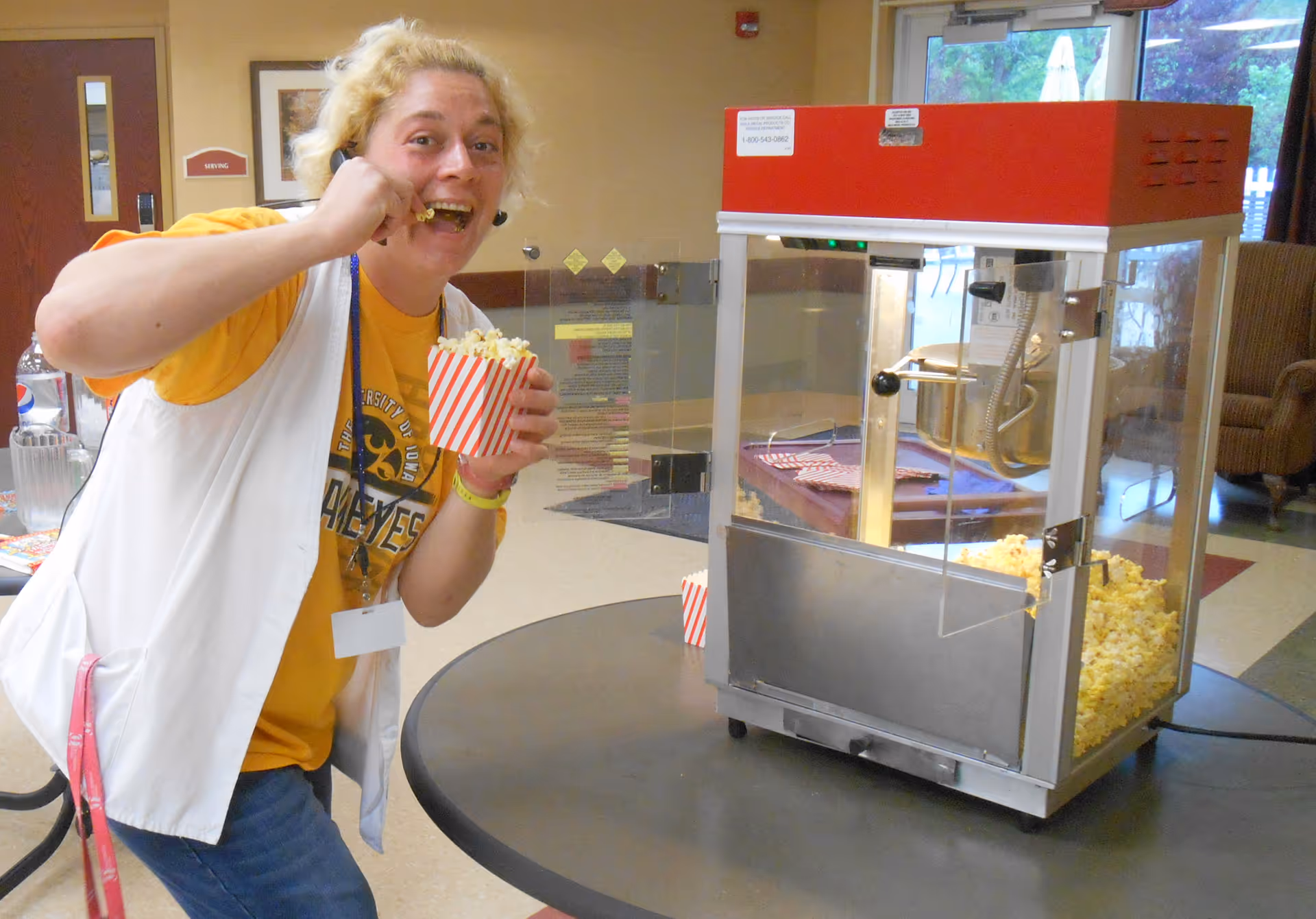 A person wearing a yellow shirt and white vest is eating popcorn from a red and white striped container next to a popcorn machine on a round table inside a room with beige walls and a glass door in the background.