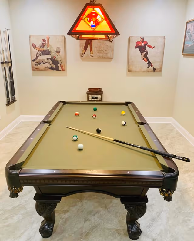 A billiards room with a pool table featuring a green felt surface, several billiard balls, and a cue stick resting on the table. The room has beige walls adorned with three vintage-style sports paintings depicting football players. A stained glass pendant light hangs above the pool table, and there is a small cabinet against the back wall.