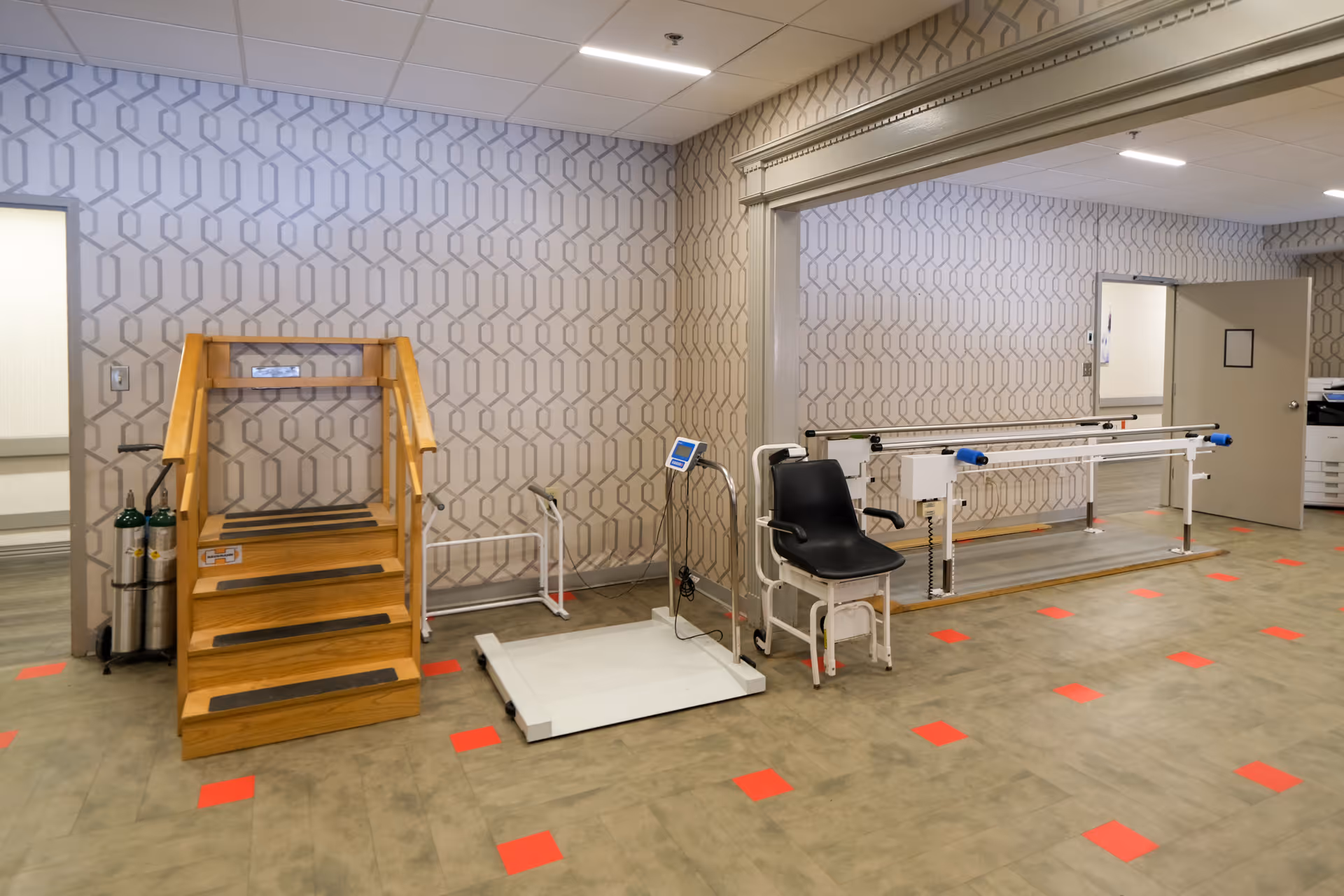 Rehabilitation room with wooden practice stairs, parallel bars, a transfer chair, scale platform, and oxygen tanks against patterned wallpaper.
