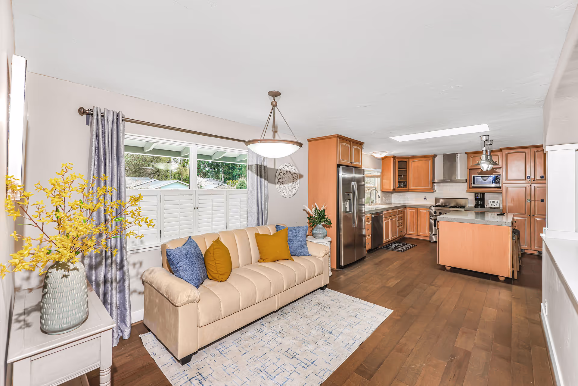 Bright and spacious interior featuring a beige sofa with blue and mustard yellow pillows, a small side table with a vase of yellow flowers, large window with gray curtains and white shutters, and an open kitchen with wooden cabinets, stainless steel appliances, and an island with bar stools.