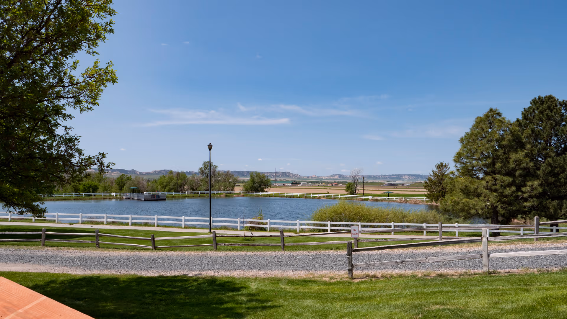A tranquil pond surrounded by white fencing, grassy lawns, trees and a gravel path under a clear blue sky.