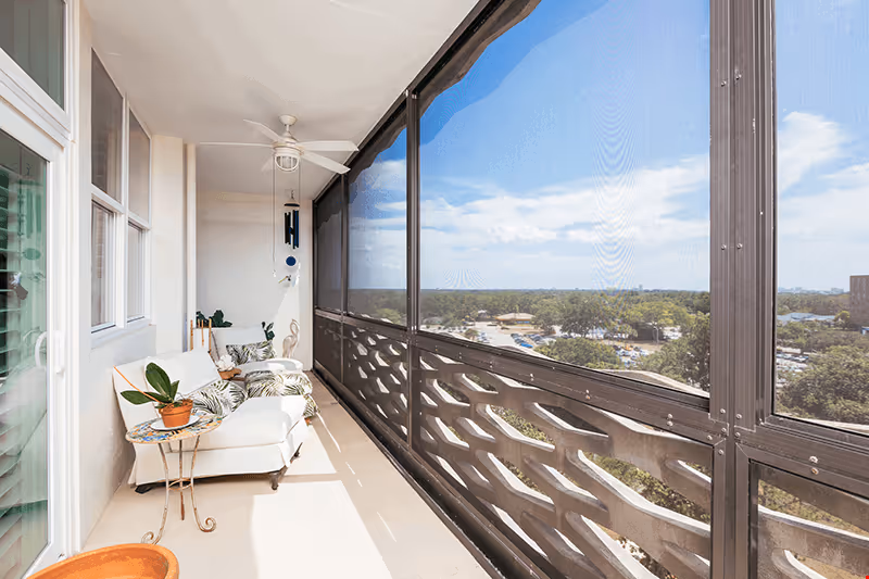 Screened-in balcony with cushioned lounge chair, side tables, potted plants and a ceiling fan overlooking trees and a distant parking area.