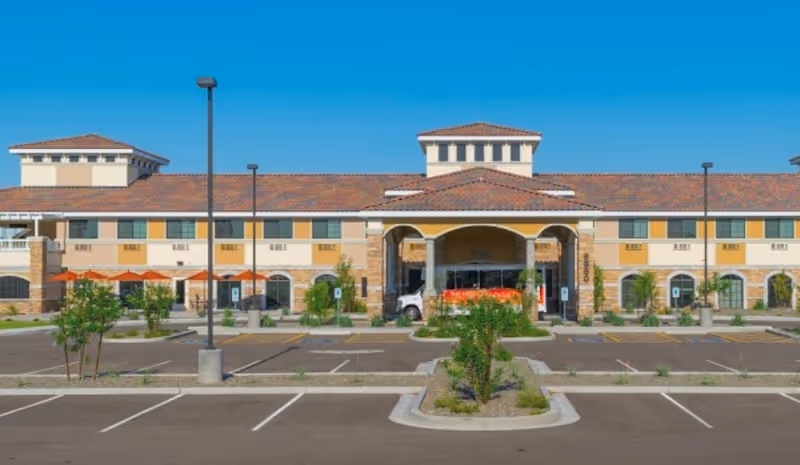 Front exterior of a two-story senior living facility with a covered entrance, parking lot, and desert landscaping under a clear blue sky.
