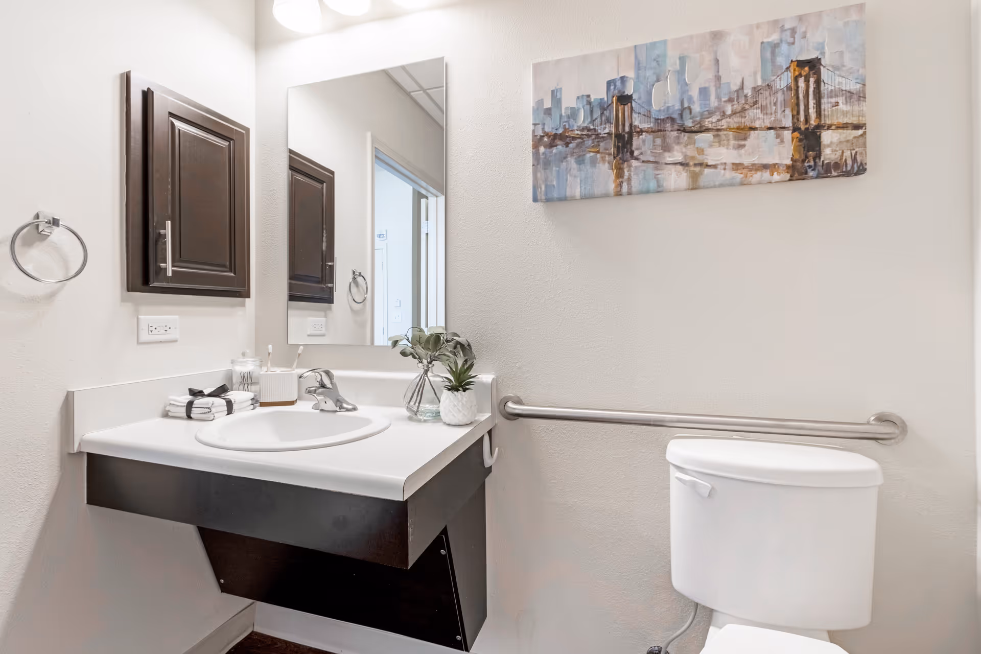 A clean and modern bathroom with a white sink and countertop, a dark wood cabinet mounted on the wall, a large mirror above the sink, a white toilet with a stainless steel grab bar on the wall behind it, and a colorful painting of a bridge hanging above the grab bar.
