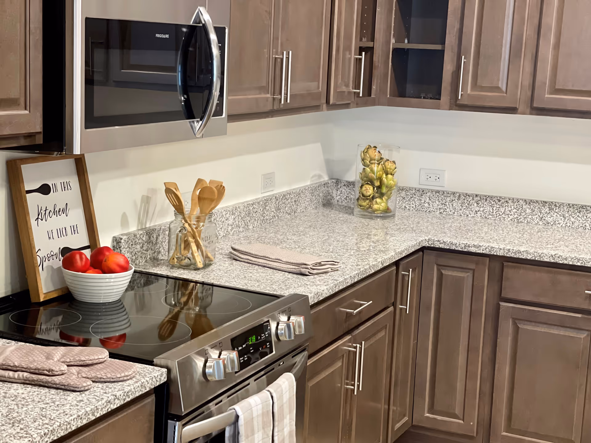 Modern kitchen corner with granite countertops, brown wooden cabinets, a stainless steel microwave and stove, a bowl of red apples, wooden cooking utensils in a glass jar, a folded kitchen towel, and a glass vase filled with artichokes.