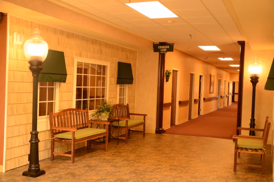 Warmly lit senior living hallway with wooden benches, streetlamp-style lamps, faux windows and a long corridor of resident rooms.