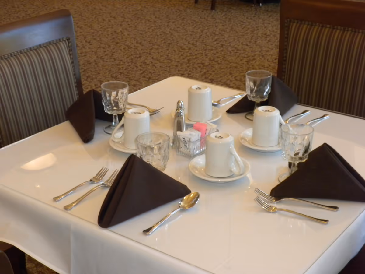 Square dining table set for four with dark folded napkins, cups, glasses, silverware and a sugar caddy.