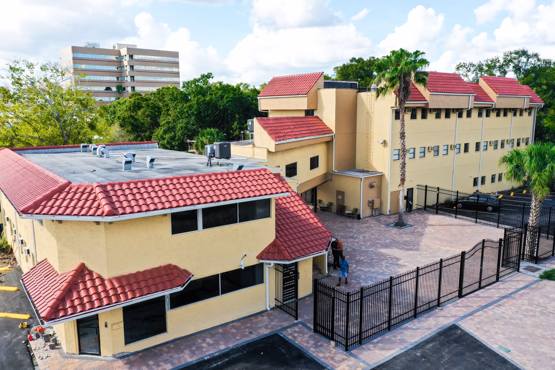 Exterior view of a senior living facility with beige walls and red tiled roofs, surrounded by a black metal fence. There are a few palm trees and a paved courtyard area with some chairs. Two people are standing near the entrance. A multi-story office building and green trees are visible in the background under a partly cloudy sky.