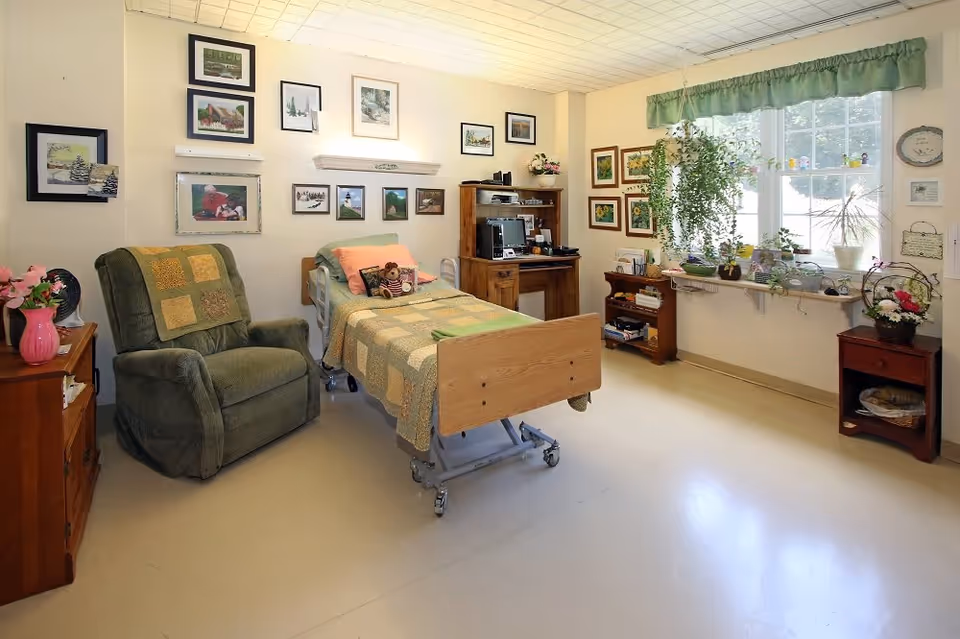 A cozy and well-lit bedroom in a senior living facility featuring a single hospital-style bed with a patchwork quilt, a green recliner chair with a matching quilt, wooden furniture including a desk with a computer and a small bookshelf, and a window with green valance and several potted plants on the windowsill. The walls are decorated with multiple framed pictures and paintings.