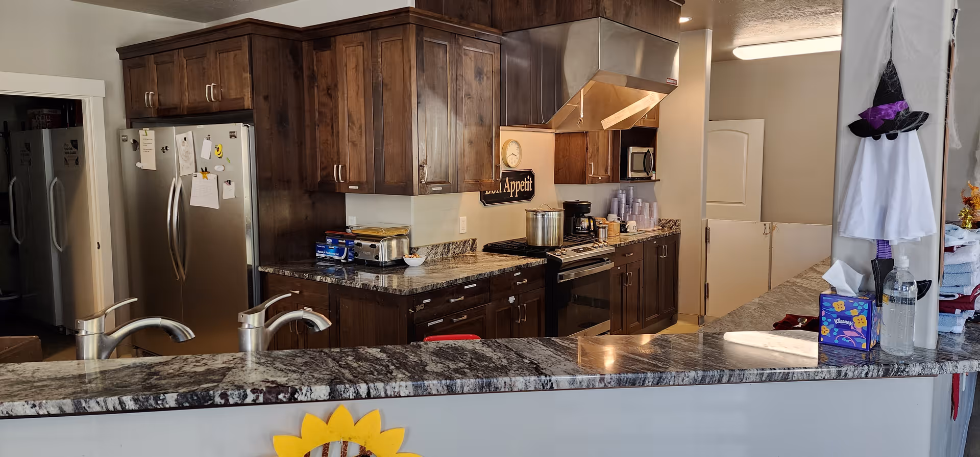 Interior communal kitchen with granite countertops, stainless steel refrigerator and stove, dark wood cabinets, and a pass-through counter.