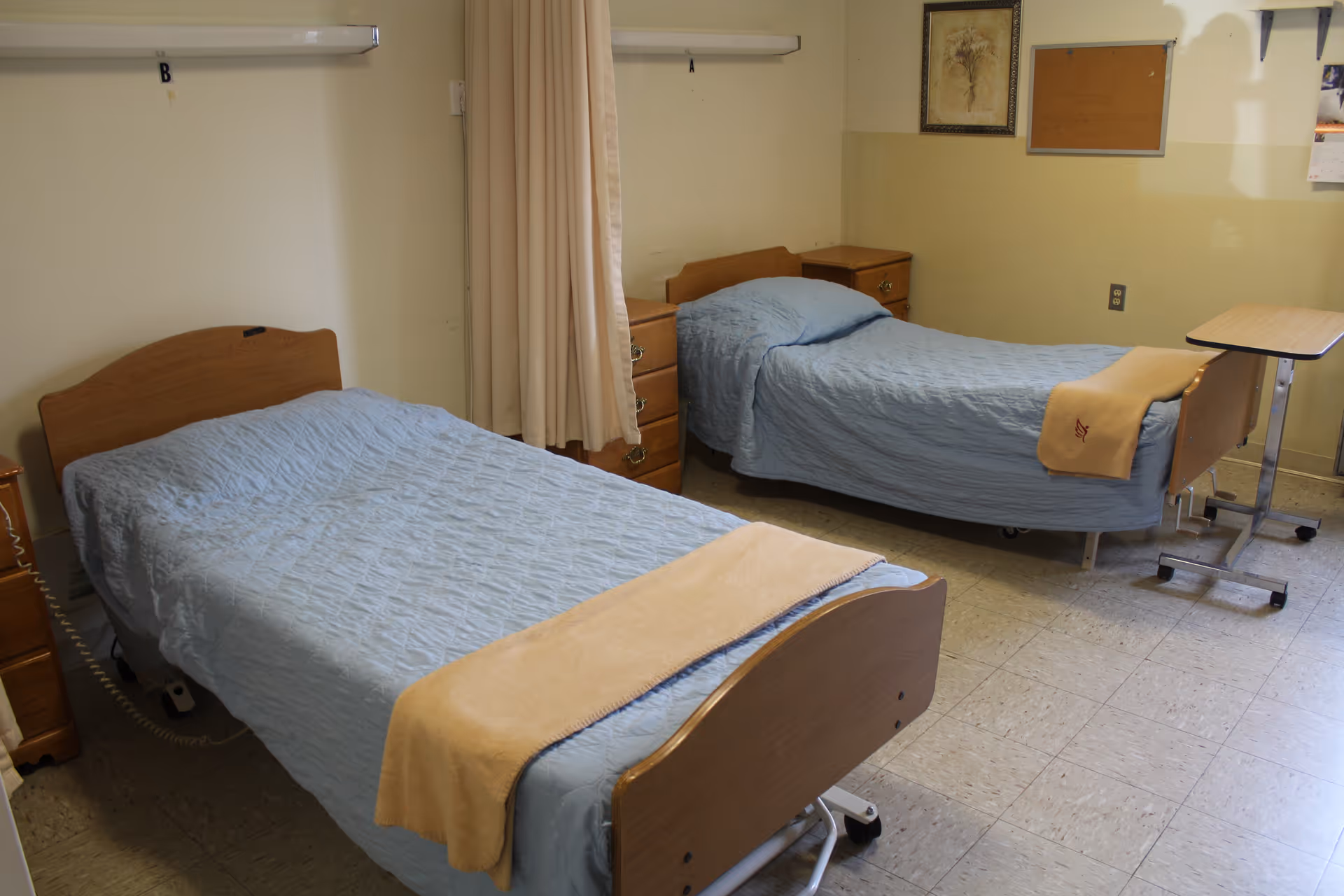 A therapy room with two hospital-style beds covered in light blue bedspreads and beige blankets at the foot. Each bed has a wooden headboard and footboard, with a small wooden nightstand between them. There is a beige curtain hanging between the beds, and a small overbed table on wheels next to one bed. The room has light-colored walls with a framed picture and a corkboard on the wall.