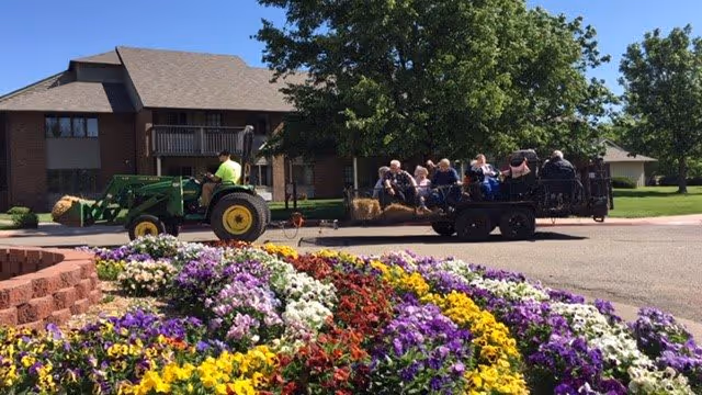 A tractor pulls a trailer of seated residents past a brick building with a colorful flowerbed in the foreground.