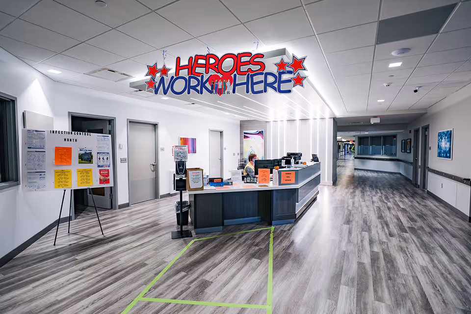 A clean and modern nursing facility hallway with a reception desk staffed by a person wearing a mask. Above the desk, a colorful sign reads 'HEROES WORK HERE' with red stars and a heart symbol. To the left, there is a bulletin board with various notices about infection control. The hallway has wood-patterned flooring, white walls, and several closed doors along the corridor.