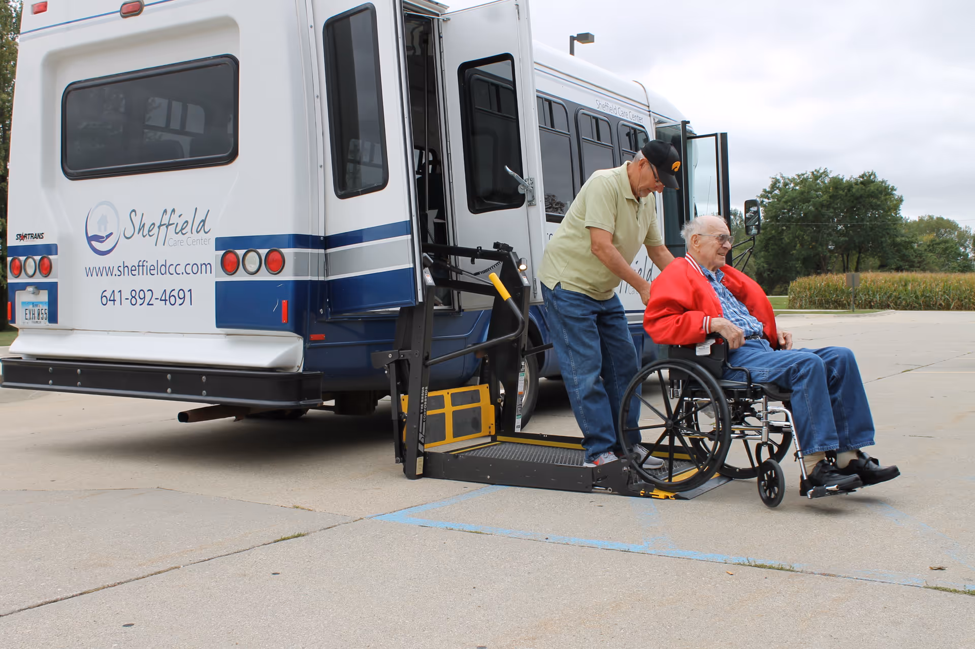 An elderly man in a wheelchair is being assisted by another man as they use a wheelchair lift to get off a Sheffield Care Center transport bus in an outdoor parking area.
