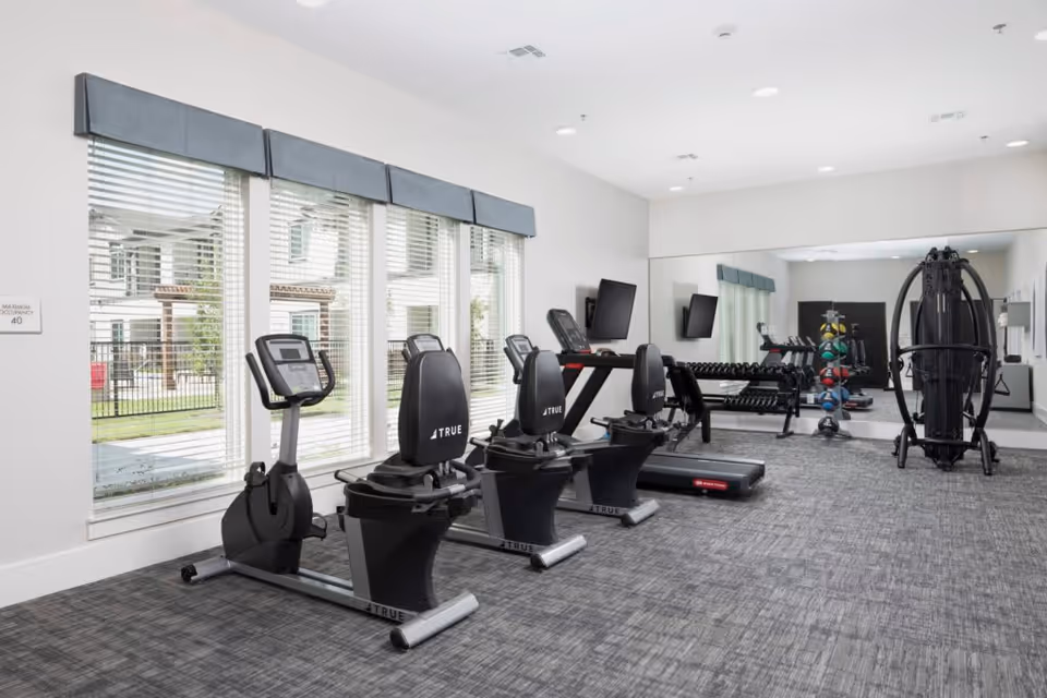 A bright fitness room with large windows covered by blinds, featuring exercise equipment including stationary bikes, a treadmill, a weight rack with dumbbells, a multi-functional gym machine, and two wall-mounted televisions. The room has gray carpet flooring and a large mirror on one wall.