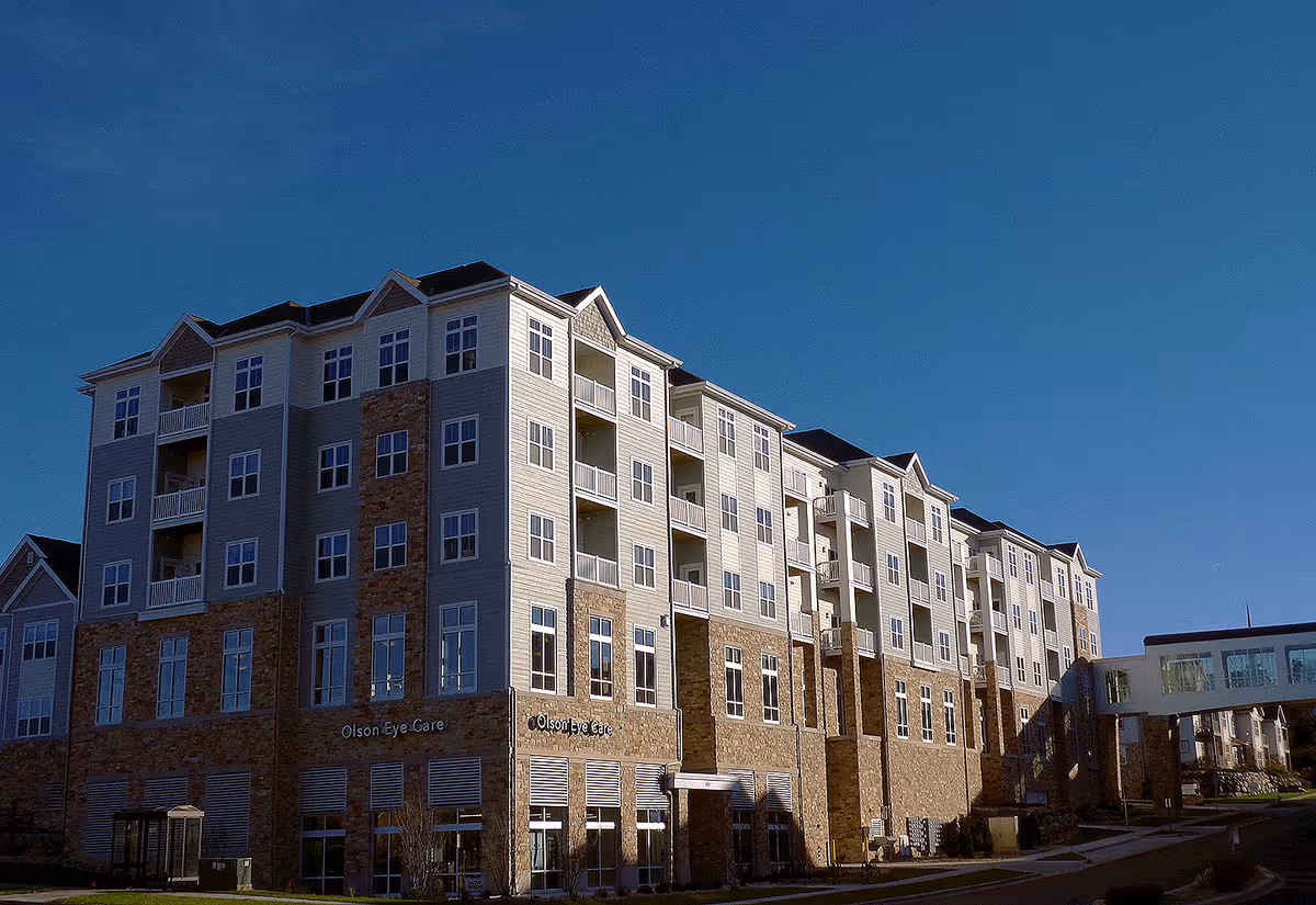 Exterior view of a multi-story residential building with a combination of stone and light-colored siding, featuring multiple windows and balconies under a clear blue sky.