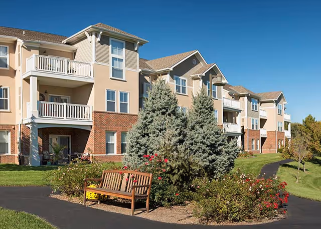 Exterior view of a multi-story senior living building with balconies, landscaped grounds, a bench, and pathways under a clear blue sky.