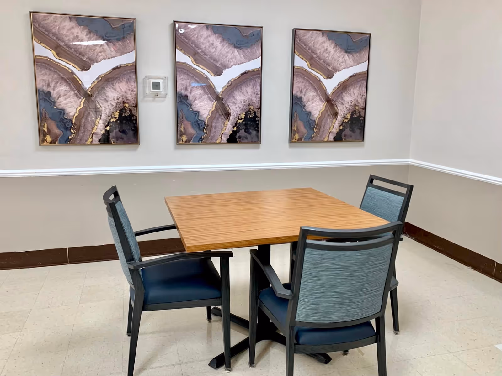 A small square wooden table with four chairs around it in a room with beige walls. Three abstract art paintings with purple, blue, and gold tones hang on the wall above the table. A thermostat is mounted on the wall between the paintings.