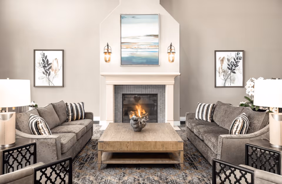 A cozy living room with two gray sofas facing each other, each adorned with striped and patterned throw pillows. Between them is a wooden coffee table with a decorative centerpiece. The room features a fireplace with a fire burning, flanked by two wall-mounted lantern-style lights. Above the fireplace hangs a landscape painting with blue and beige tones. Two framed botanical prints are on the walls beside the fireplace. The room is softly lit by table lamps on side tables next to each sofa.