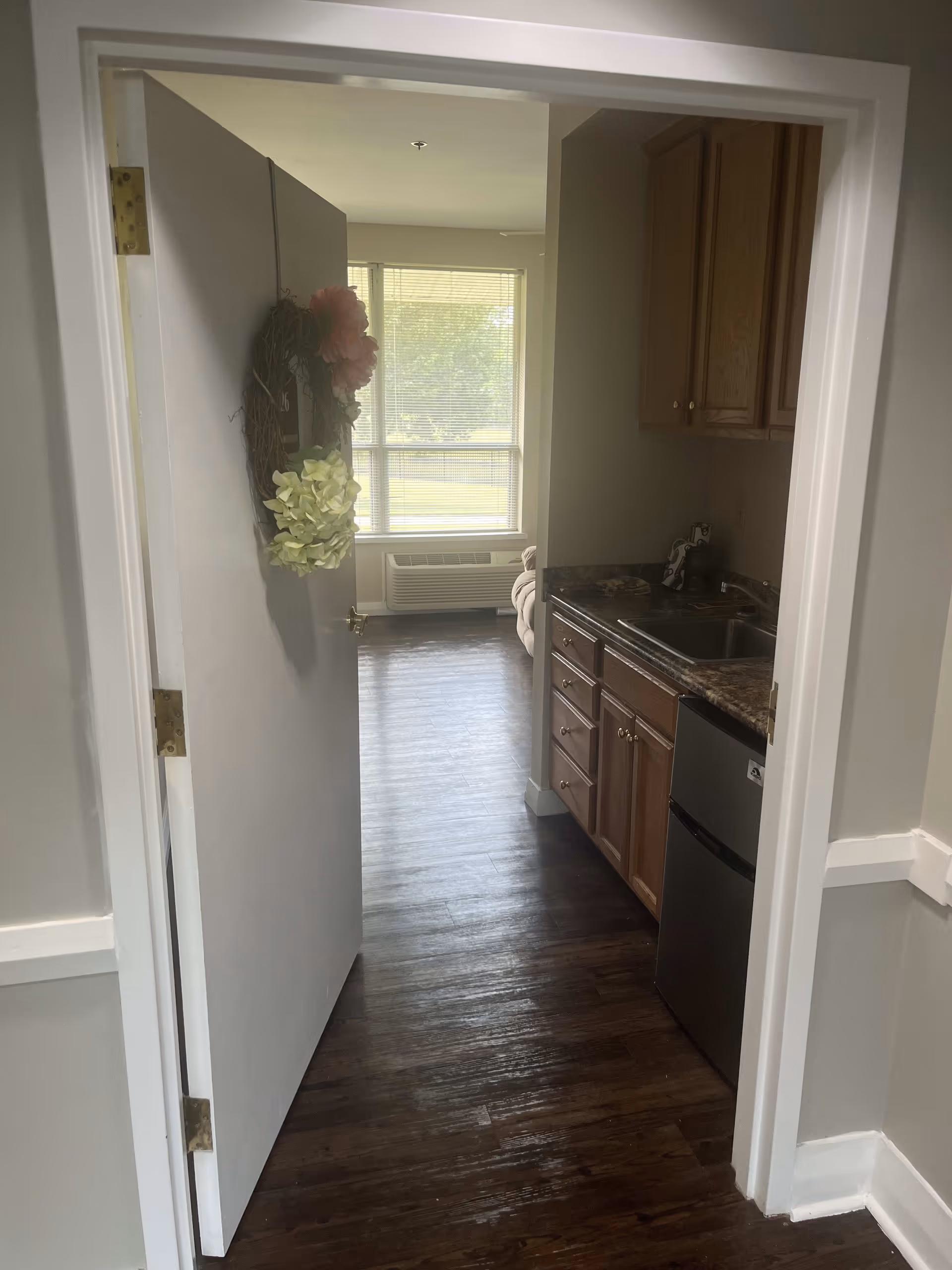 View through an open door into a small kitchen area with wooden cabinets, a countertop with a sink, and a mini refrigerator. Beyond the kitchen, there is a living room area with a large window letting in natural light and a beige couch partially visible. The floor is dark wood, and the door has a decorative wreath with flowers.