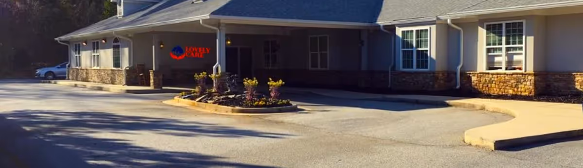 Exterior view of a single-story building with stone and beige siding, featuring multiple windows and a covered entrance with a small landscaped area in front. A car is parked on the left side. The building has a sign that reads 'Lovely Care' in red and blue near the entrance.