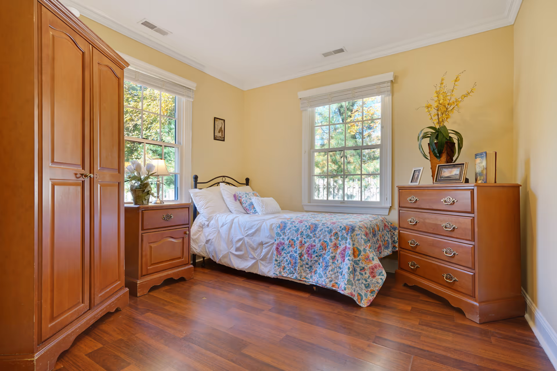A cozy bedroom with a single bed covered in white bedding and a colorful floral quilt. The room has two large windows with white blinds, wooden furniture including a wardrobe, a nightstand with a lamp and flowers, and a chest of drawers with framed pictures and a potted plant. The walls are painted light yellow and the floor is wooden.