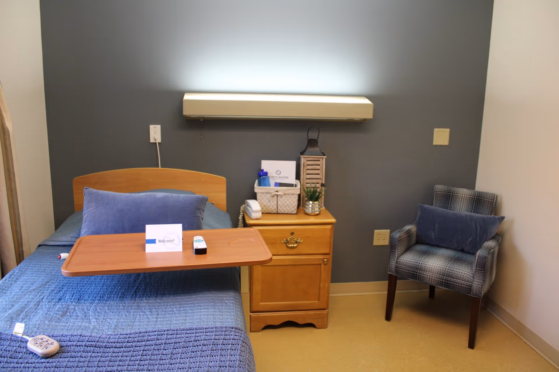 A neatly made single bed with an overbed tray, wooden nightstand and upholstered chair against a gray wall.
