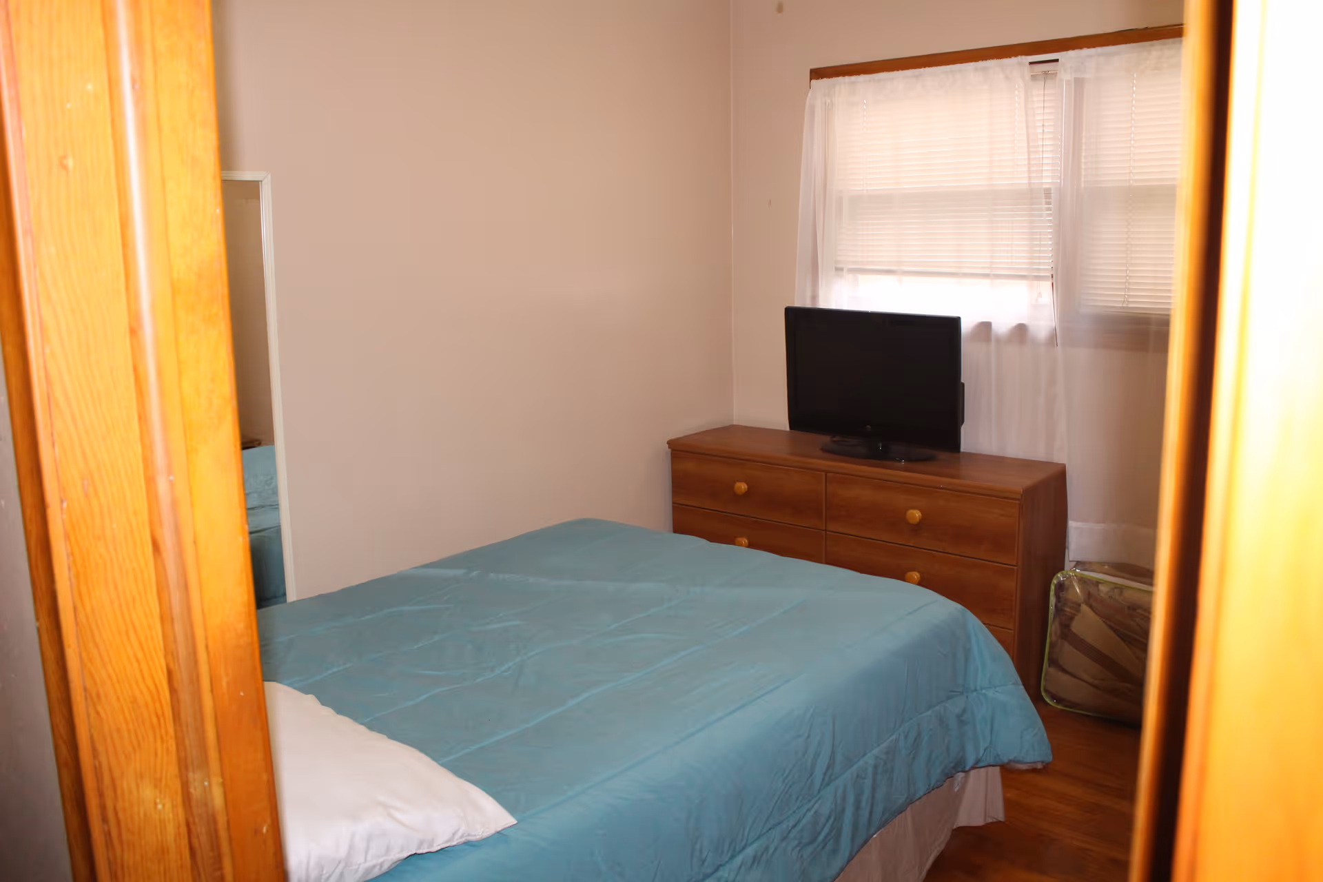 Small bedroom with a teal bedspread, wooden dresser topped by a TV, and a window with blinds.