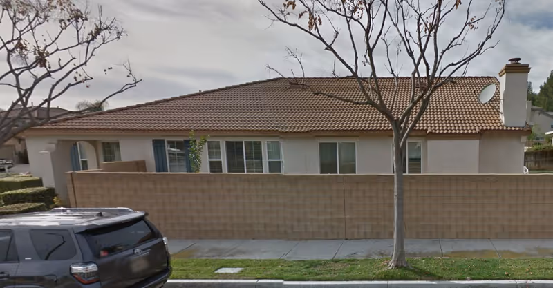 Single-story residential building with a tiled roof behind a beige brick wall, a tree with no leaves in front, and a black SUV parked on the street.