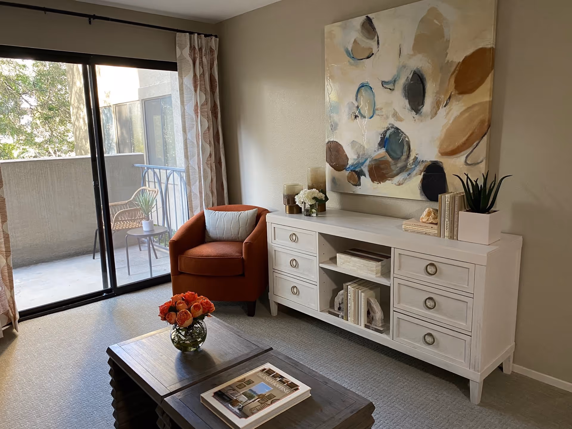 A cozy living room area with a sliding glass door leading to a balcony. The room features a burnt orange armchair with a cushion, a white sideboard with decorative items including books, plants, and candles, and a wooden coffee table with a vase of orange roses and a book. A large abstract painting hangs on the wall above the sideboard.