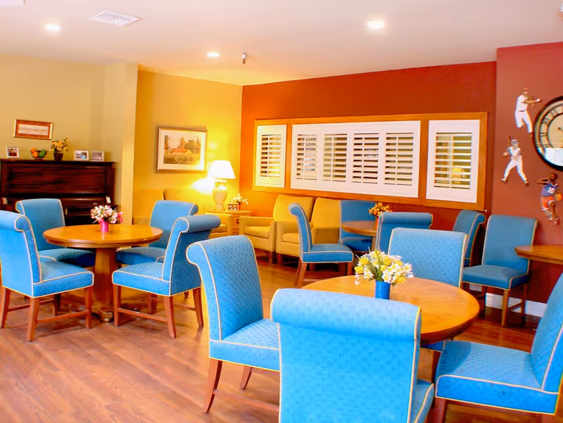 A cozy common area with round wooden tables surrounded by blue upholstered chairs. The room has warm lighting, a wooden floor, a piano against one wall, framed pictures, a table lamp, and decorative wall art including a large clock and sports figures.