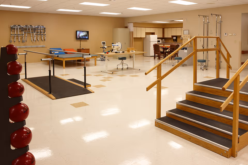 A spacious physical therapy room with equipment including parallel bars, a set of stairs with handrails, therapy tables, exercise balls, walkers mounted on the wall, and a small kitchen area with a refrigerator and chairs in the background.