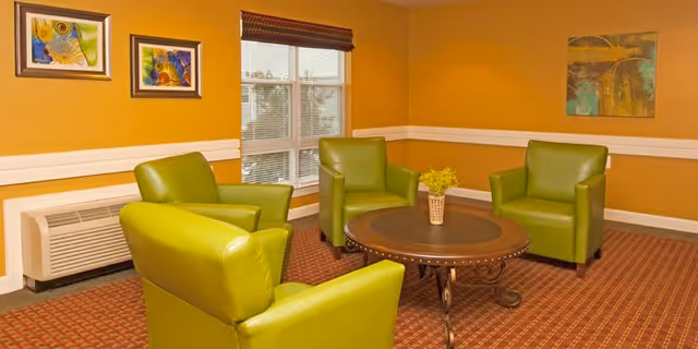 A cozy sitting area with four green armchairs arranged around a round wooden table with a small plant centerpiece. The room has mustard yellow walls with white trim, a window with blinds, and three colorful paintings hanging on the walls.