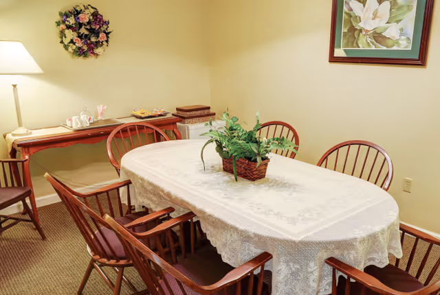 A dining room with a rectangular table covered with a white lace tablecloth and a green potted plant centerpiece. Six wooden chairs with armrests surround the table. Against the wall is a wooden side table with a lamp, a floral wreath hanging above it, and some items placed on the table. A framed floral painting hangs on the adjacent wall.